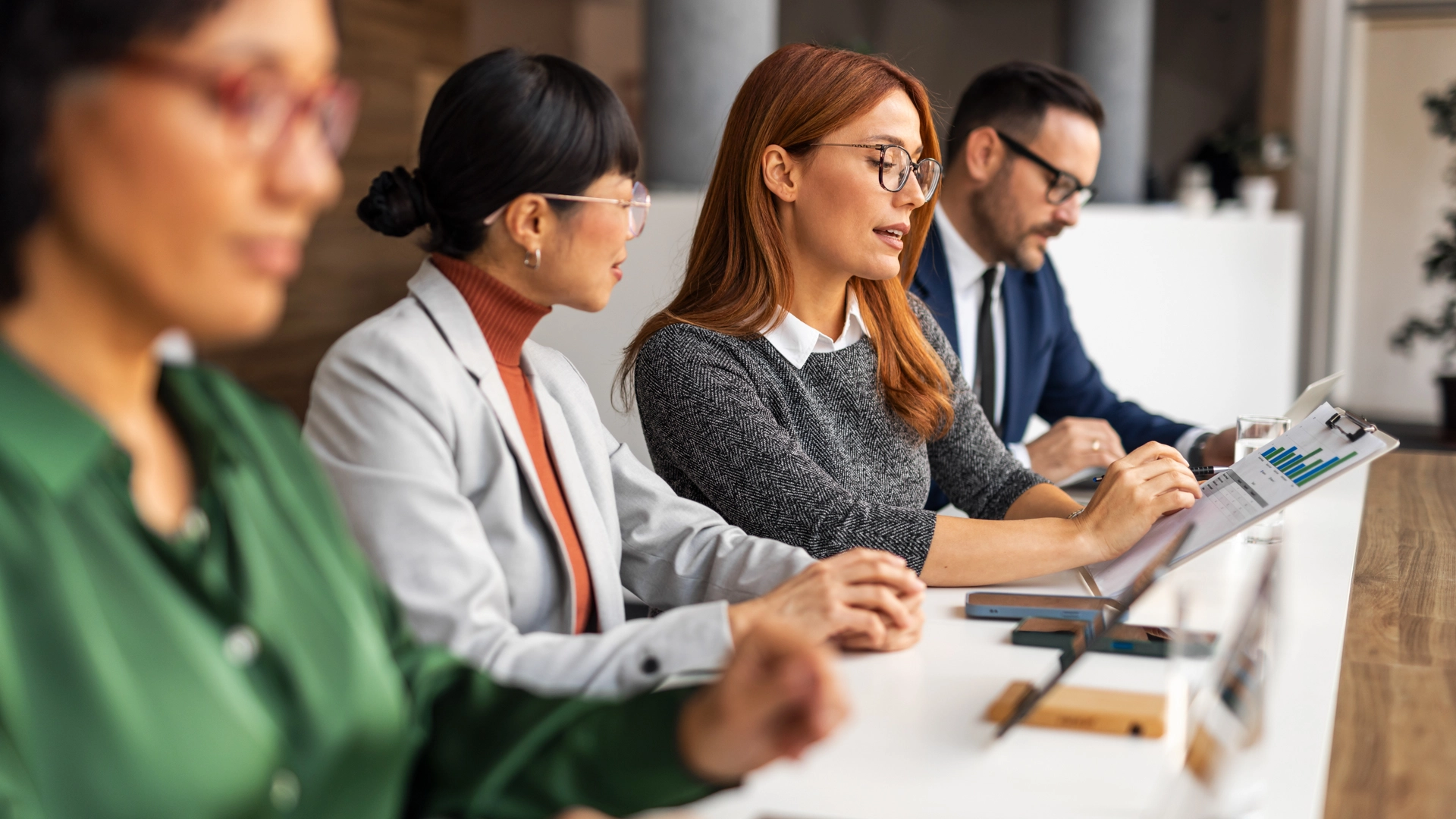 A group of diverse professionals sit at a long white table in a bright office; a woman with red hair and glasses points to a bar chart on a clipboard while discussing data with her colleagues.