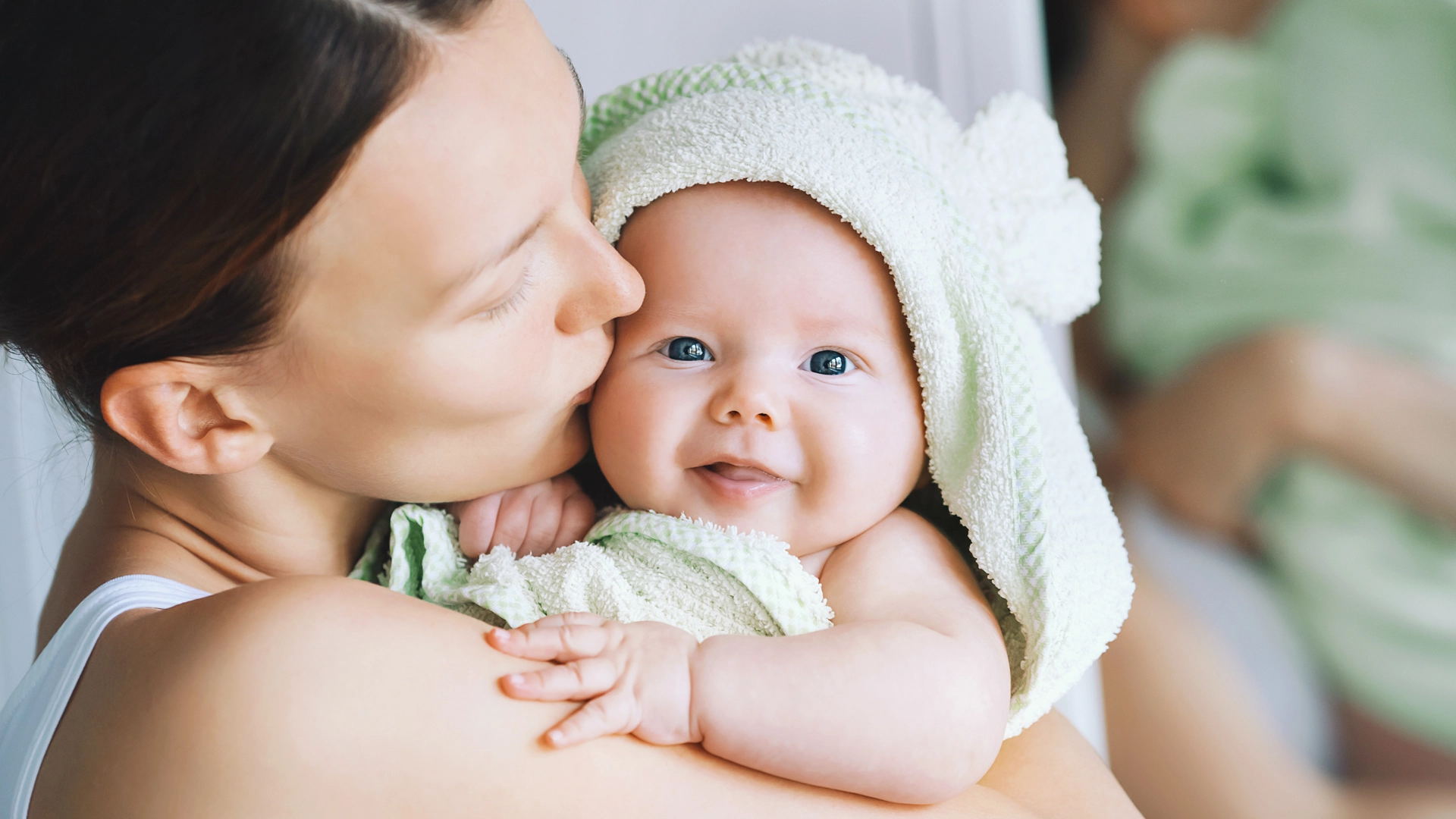 A mother tenderly kisses the cheek of her smiling infant, who is wrapped in a soft, light green hooded towel.