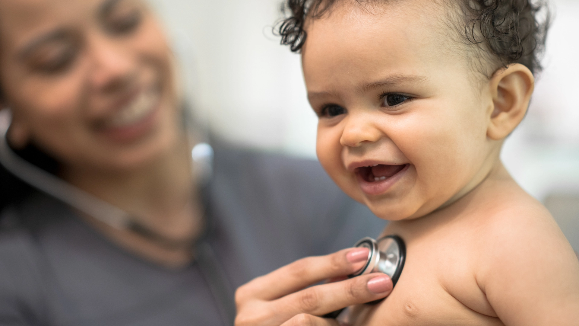 A close-up of a happy baby smiling while a healthcare provider uses a stethoscope to listen to their heartbeat.