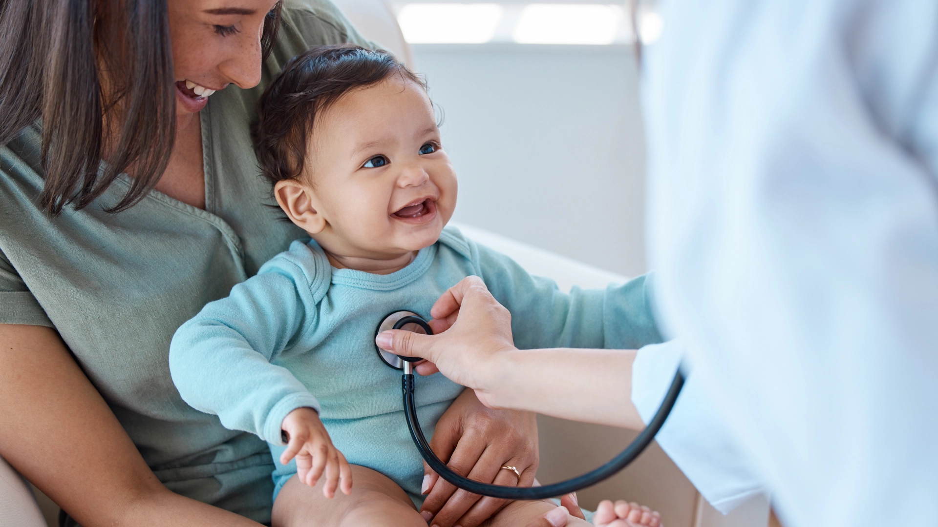 A healthcare professional uses a stethoscope to examine a smiling baby who is being held securely in its mother's lap.
