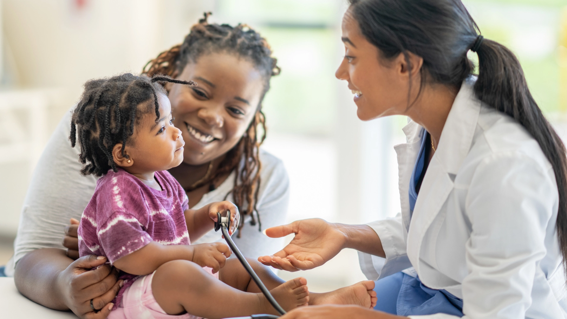 A pediatrician interacts playfully with a young child sitting on their mother's lap, showing the child how a stethoscope works.