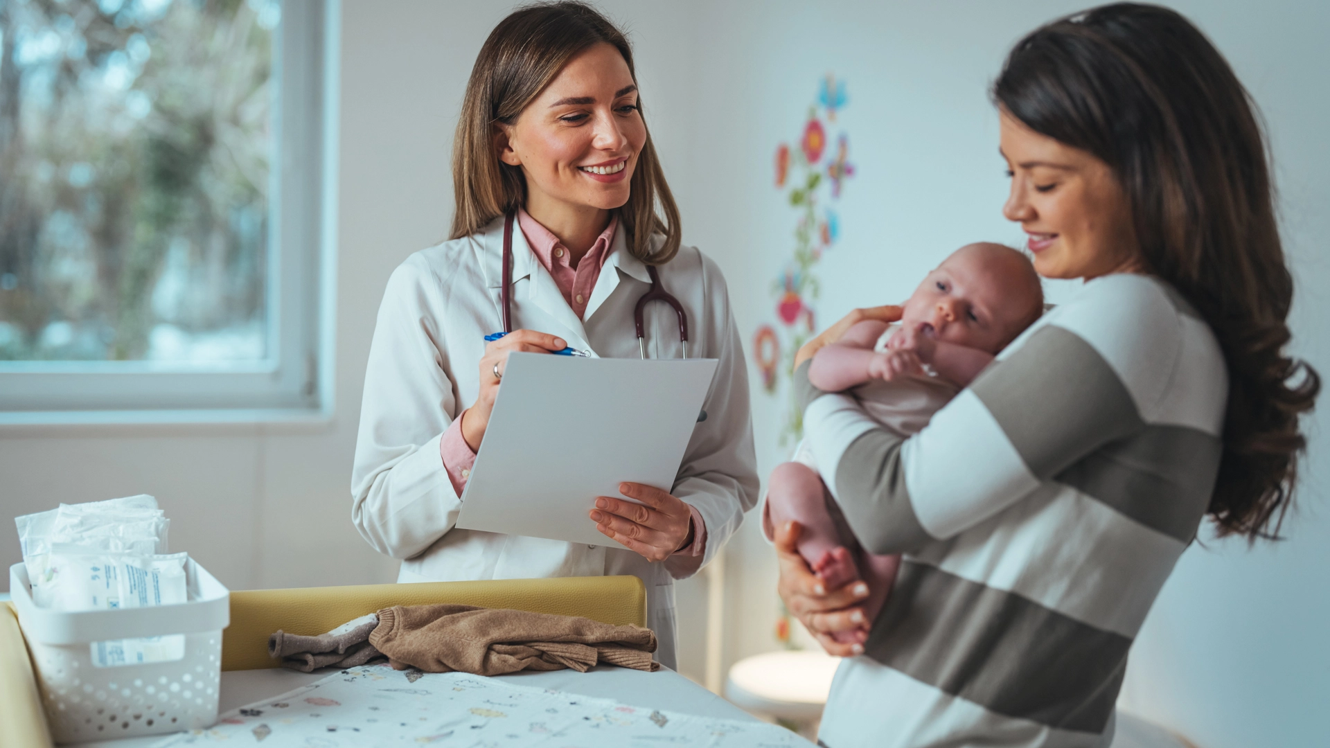 A female doctor in a white coat smiles while writing on a clipboard, as a mother holds her newborn baby nearby in a clinical setting.